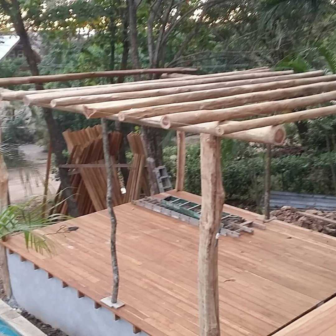 Rafters laid across the rancho posts before the thatch goes on, jungle behind.
