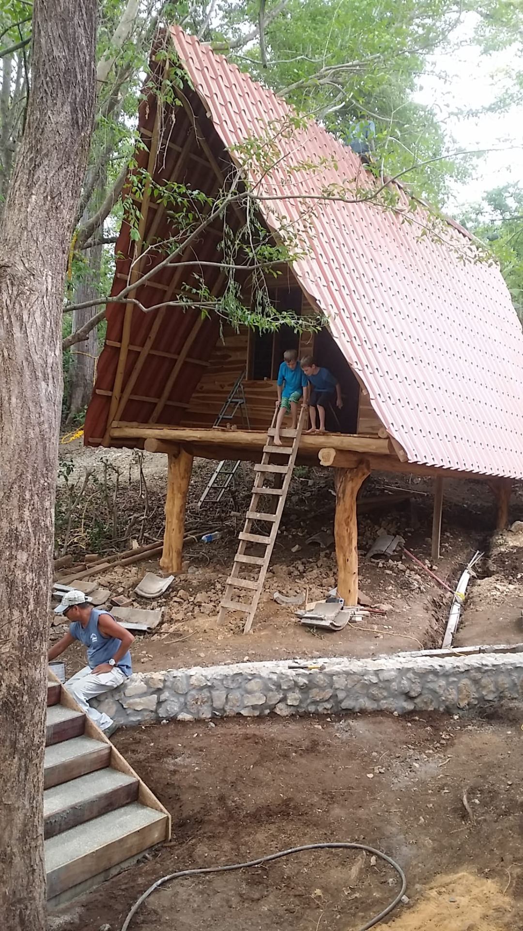 Treehouse Chilo nearly complete, red clay-tile roof on the A-frame, two kids peeking out of the loft opening.